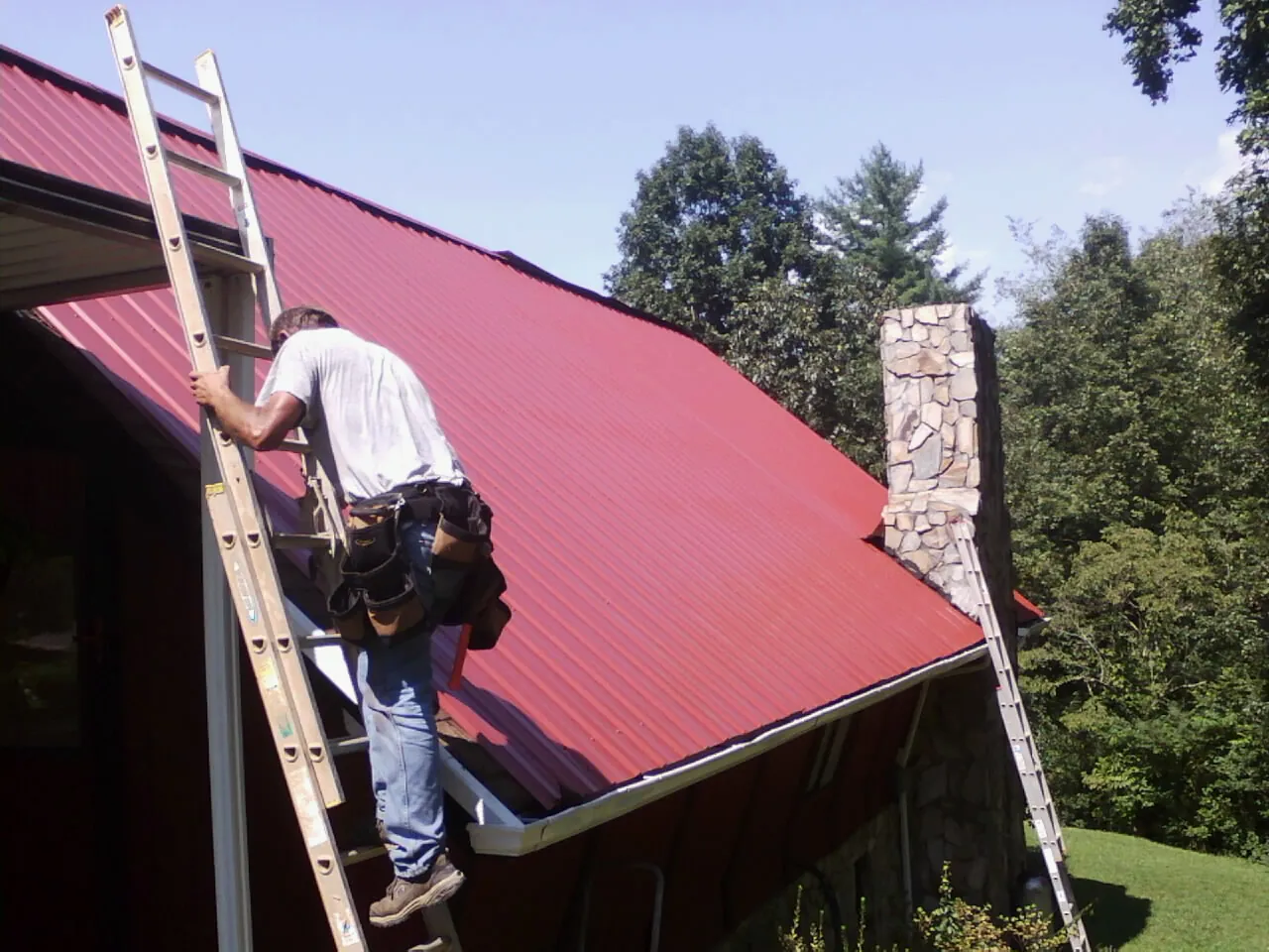 Roofer on a ladder beside a red metal roof and stone chimney — Soffit & Fascia Repair by Roofing Brothers in Glen Carbon